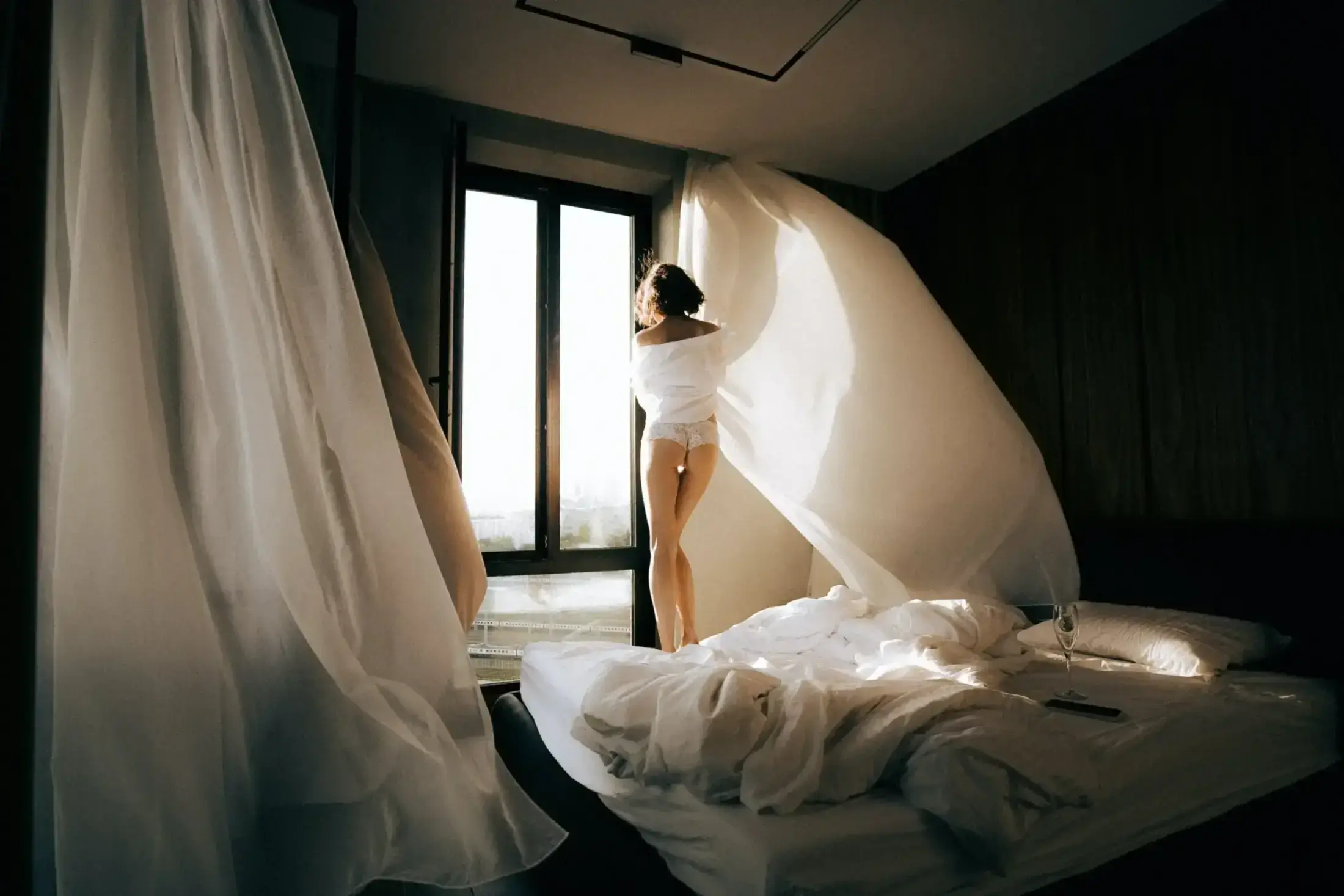 Woman standing by bedroom window with billowing white curtains.